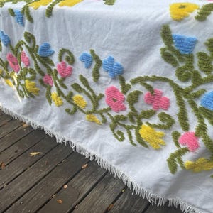 May include: White crocheted throw blanket with a floral design featuring pink, yellow, blue, and green flowers. The blanket has a fringe border.