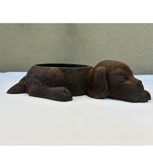 May include: A dark brown, cast iron planter shaped like a sleeping dog. The dog is lying down with its head resting on its front paws, and the circular opening for a plant is located in the center of the dog's back, featuring a textured finish.