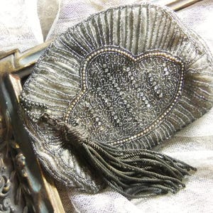 May include: An antique, silver-toned beaded purse with a heart-shaped design. The purse features a beaded tassel and is set against a textured, off-white background. The purse is approximately 6 inches wide.