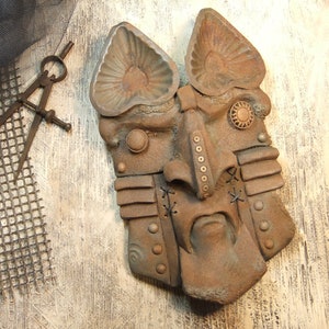 May include: A rusty metal mask with a distressed finish. The mask has a human-like face with a large nose, stitched-up eyes, and a wide mouth. The mask is decorated with metal rivets and other embellishments.