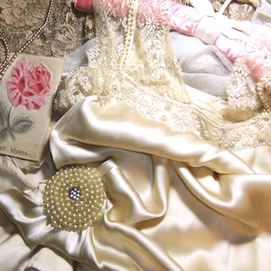 May include: A cream-colored satin camisole featuring intricate lace trim at the neckline, paired with a round pearl-encrusted brooch. A vintage-style card with a pink rose illustration and the text "BEST WISHES" rests nearby with a string of pearls.