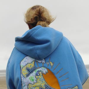 May include: A light blue hooded sweatshirt with a colorful wave and sun design on the back. The wave is made of patchwork fabric with yellow, blue, and green patterns. The sun is orange with yellow rays.