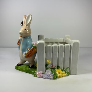 May include: A white ceramic planter shaped like a picket fence with a Peter Rabbit figurine holding a shovel. The planter has a green base with colorful flowers.