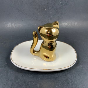 May include: A gold-colored cat figurine sits atop a white, oval-shaped dish with a gold trim. The cat is posed with its tail curved upward. The dish is a decorative item, possibly for jewelry or small items.