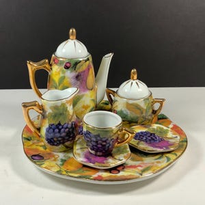 May include: A porcelain tea set featuring a teapot, creamer, sugar bowl, and a cup with a saucer, all arranged on a matching serving tray. The pieces are decorated with a colorful fruit pattern including purple grapes and gold-toned trim.