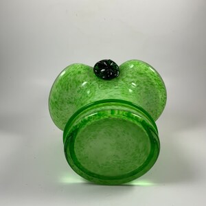 Op de afbeelding: Een groene glazen vaas met een gespikkeld patroon en een donkergroen, bloemvormig accent aan de bovenkant. De vaas heeft een brede, afgeronde vorm met een uitlopende rand en een cirkelvormige basis. Het glas is doorschijnend.