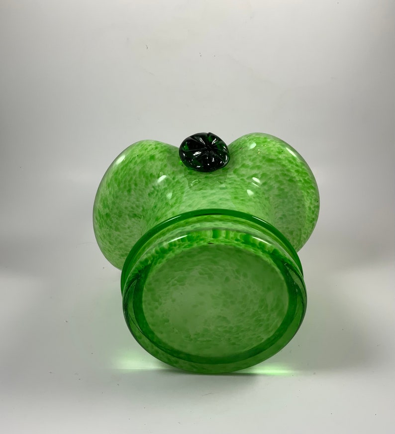 Op de afbeelding: Een groene glazen vaas met een gespikkeld patroon en een donkergroen, bloemvormig accent aan de bovenkant. De vaas heeft een brede, afgeronde vorm met een uitlopende rand en een cirkelvormige basis. Het glas is doorschijnend.