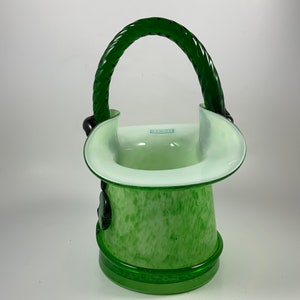 Op de afbeelding: Een groene glazen vaas met een handvat. De vaas heeft een gespikkeld patroon en een witte rand. Het handvat is een gedraaid ontwerp in een donkerdere groene kleur. De basis is effen groen.