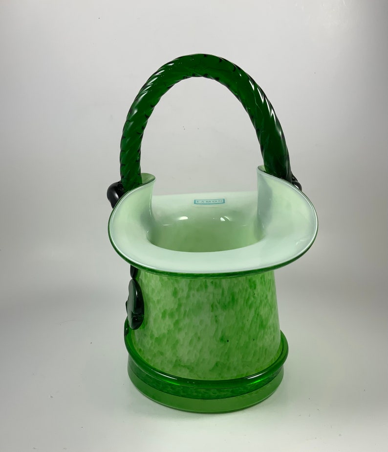 Op de afbeelding: Een groene glazen vaas met een handvat. De vaas heeft een gespikkeld patroon en een witte rand. Het handvat is een gedraaid ontwerp in een donkerdere groene kleur. De basis is effen groen.