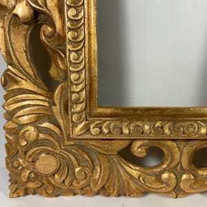 May include: An ornate gold picture frame with detailed carvings. The frame has a rectangular opening and a heavily textured surface with a distressed finish. The design includes scrolling leaves and floral motifs.