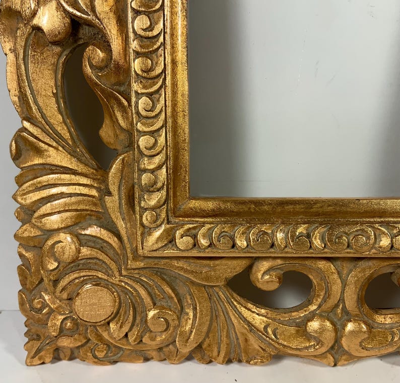 May include: An ornate gold picture frame with detailed carvings. The frame has a rectangular opening and a heavily textured surface with a distressed finish. The design includes scrolling leaves and floral motifs.