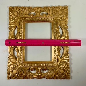 May include: An ornate gold picture frame with detailed scrollwork and a rectangular aperture. A pink ruler is placed across the frame, measuring approximately 30 cm in length, indicating the frame's size.