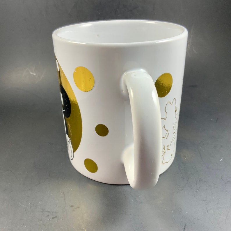 May include: White ceramic mug with a handle, featuring gold polka dots and a gold silhouette of a cartoon character. The mug has a glossy finish and is set against a gray background.