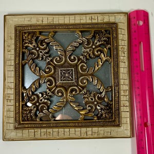 May include: Square decorative wall art with a cream-colored mosaic border and a gold-toned filigree design. The center features a light blue background. The pink ruler next to the art measures up to 12 inches.