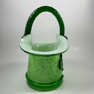 Op de afbeelding: Een groene glazen vaas met een handvat. De vaas heeft een gespikkeld patroon en een witte rand. Het handvat is een gedraaid ontwerp. De vaas heeft een ronde basis en is ongeveer 20 cm hoog.