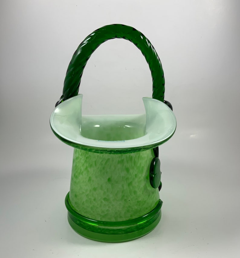 Op de afbeelding: Een groene glazen vaas met een handvat. De vaas heeft een gespikkeld patroon en een witte rand. Het handvat is een gedraaid ontwerp. De vaas heeft een ronde basis en is ongeveer 20 cm hoog.