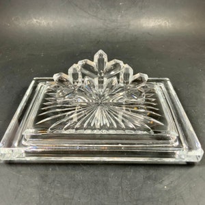 May include: A clear, rectangular glass butter dish with a decorative, starburst pattern. The lid features a stylized, floral-like design. The dish is made of thick, clear glass and sits on a dark surface.