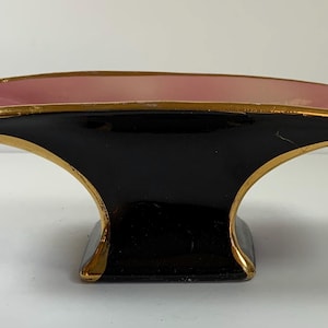 May include: A decorative, art deco-style dish with a black base and a pink interior. The rim is accented with gold trim. The dish has a unique, curved shape, and is likely made of ceramic or porcelain.