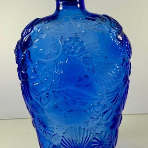 May include: A cobalt blue glass bottle with a cork stopper, featuring a raised, embossed design of various seashells, starfish, and coral patterns covering the entire exterior surface of the rounded, wide-bodied vessel.