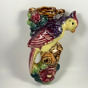 May include: A vintage ceramic wall pocket in the shape of a bird perched on a tree branch. The bird has yellow, burgundy, and purple plumage. The branch and flowers are brown, green, and burgundy. The wall pocket has a small opening at the top.