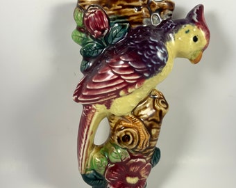 May include: A vintage ceramic wall pocket in the shape of a bird perched on a tree branch. The bird has yellow, burgundy, and purple plumage. The branch and flowers are brown, green, and burgundy. The wall pocket has a small opening at the top.