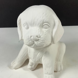 May include: A white ceramic puppy figurine in a sitting position. The unpainted sculpture features molded fur texture, floppy ears, and a detailed facial expression with visible eyes, a nose, and a mouth. The surface is matte and smooth.