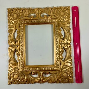 May include: Ornate gold picture frame with detailed floral and scrollwork carvings. The rectangular frame has a central opening. A bright pink ruler is placed next to it. The frame is approximately 46 cm tall.