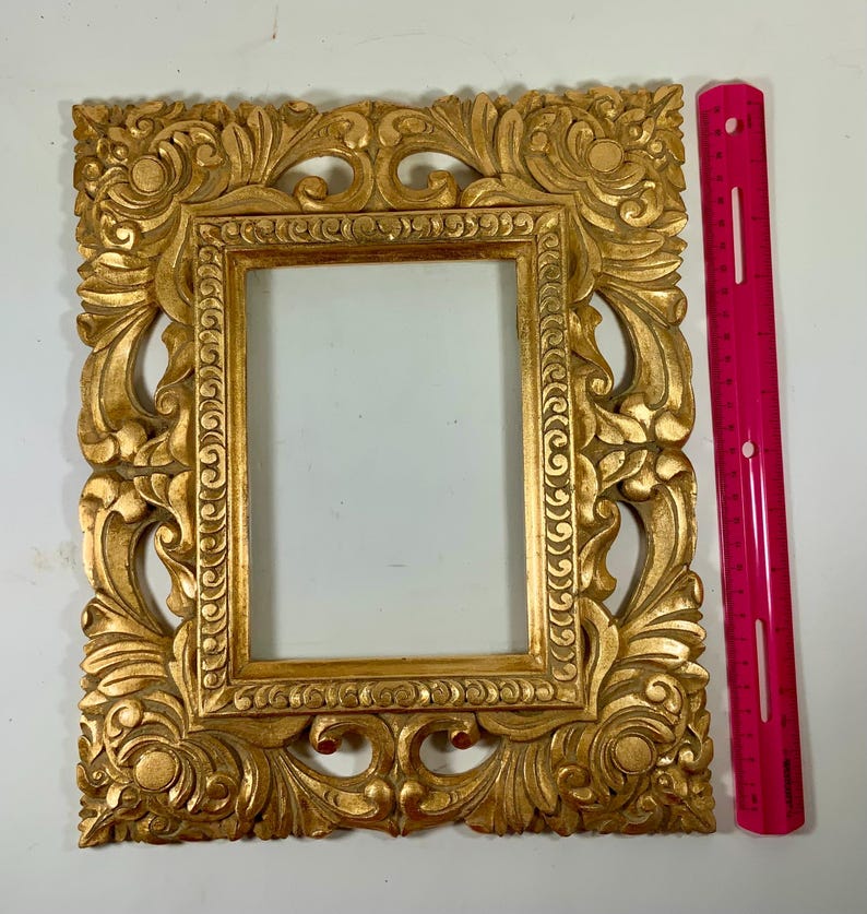 May include: Ornate gold picture frame with detailed floral and scrollwork carvings. The rectangular frame has a central opening. A bright pink ruler is placed next to it. The frame is approximately 46 cm tall.