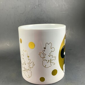 May include: White ceramic mug featuring gold polka dots and gold silhouettes of a cartoon character. The mug has a gold and black design on the side. The mug is cylindrical and has a smooth, glossy finish.