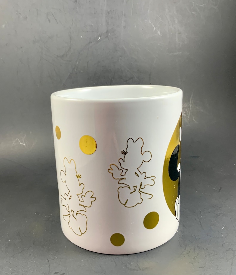 May include: White ceramic mug featuring gold polka dots and gold silhouettes of a cartoon character. The mug has a gold and black design on the side. The mug is cylindrical and has a smooth, glossy finish.