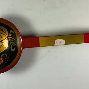 May include: A wooden ladle featuring a round bowl painted black with gold, orange, and green floral and berry motifs. The handle is painted red with a central gold band and a small white rectangular sticker attached to the middle of the handle.