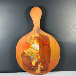 May include: A wooden cutting board with a hand-painted scene of a doll sitting in a barrel with a patchwork quilt. A small mouse is painted on the board near the barrel.