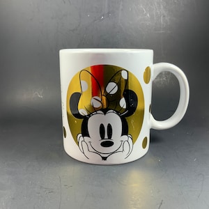May include: White ceramic mug featuring Minnie Mouse. The mug has a gold foil design with Minnie's face and a polka dot bow. Additional gold polka dots are scattered around the mug.