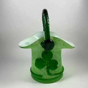 May include: A green glass basket with a woven handle and a shamrock design. The basket has a light green speckled body and a white rim. The shamrock is a darker shade of green.