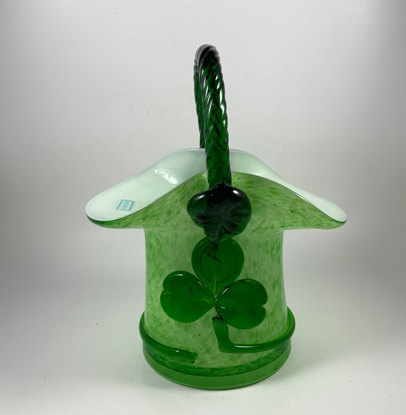 Op de afbeelding: Een groene glazen mand met een gevlochten handvat en een klaverontwerp. De mand heeft een lichtgroene gespikkelde body en een witte rand. De klaver is een donkerdere tint groen.