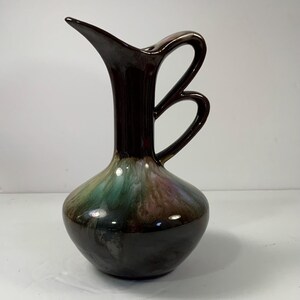 May include: A ceramic pitcher with a dark brown glaze and a unique handle design. The base of the pitcher features a blend of green, purple, and brown hues. The pitcher has a long, slender neck and a curved spout.
