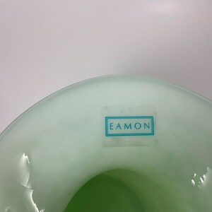 Op de afbeelding: Een lichtgroene glazen vaas met een brede, gebogen rand. De binnenkant is een donkerdere tint groen. Een rechthoekige sticker met het woord "EAMON" in een blauw kader is op de zijkant van de vaas bevestigd. De vaas staat tegen een witte achtergrond.