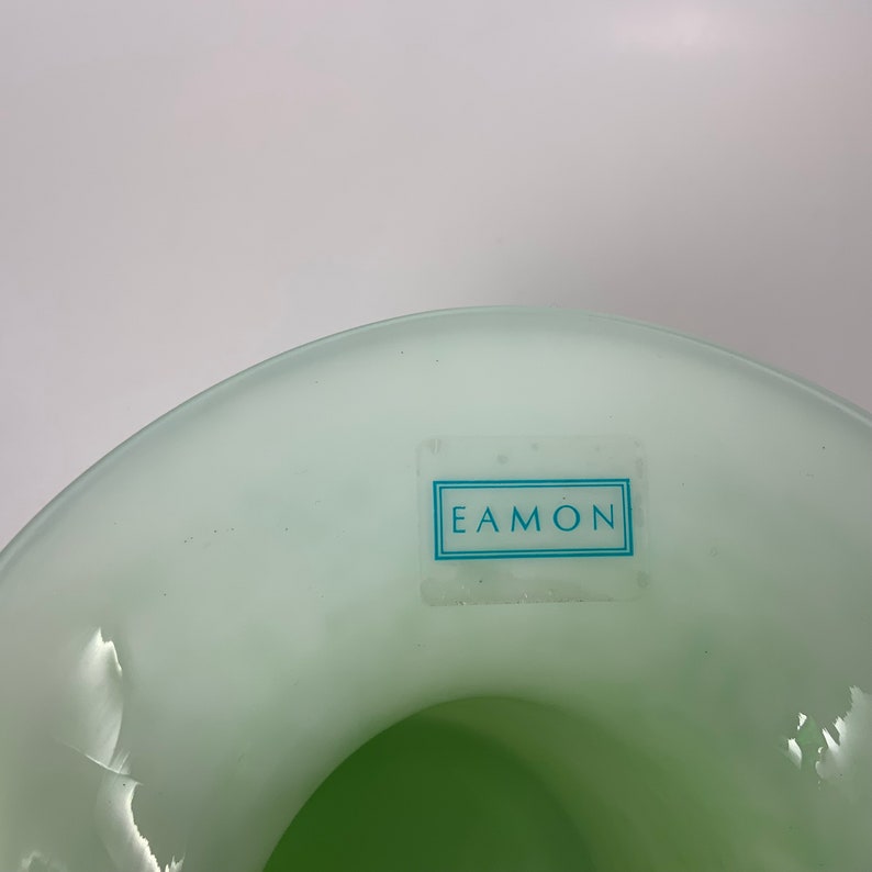 Op de afbeelding: Een lichtgroene glazen vaas met een brede, gebogen rand. De binnenkant is een donkerdere tint groen. Een rechthoekige sticker met het woord "EAMON" in een blauw kader is op de zijkant van de vaas bevestigd. De vaas staat tegen een witte achtergrond.