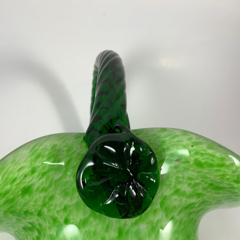 Op de afbeelding: Een groene glazen mand met een gedraaide handgreep. De mand heeft een gespikkeld patroon en een donkergroene handgreep en bodem. De mand wordt waarschijnlijk gebruikt voor decoratieve doeleinden.