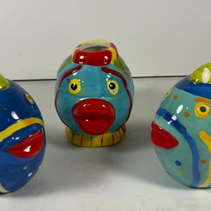 May include: Three ceramic fish figurines with colorful, abstract designs. The fish are blue, green, and yellow with red lips and white accents.