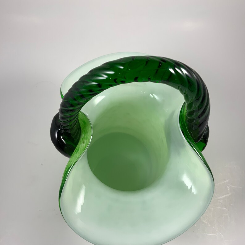 Op de afbeelding: Een groene en witte glazen mand met een gedraaide groene handgreep. De mand heeft een uitlopende rand en een afgeronde body. De handgreep is dik en heeft een touwachtige textuur. Het glas heeft een glanzende afwerking.