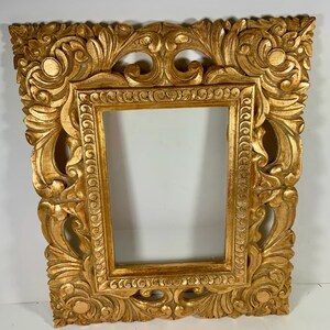 May include: An ornate gold picture frame with a rectangular aperture. The frame has detailed, carved floral and scrollwork designs. The frame is made of wood and has a distressed, antique finish. Suitable for displaying artwork or photographs. The frame measures approximately 50 cm x 40 cm.