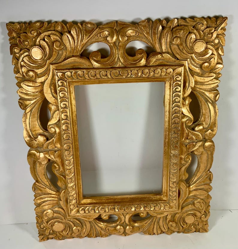 May include: An ornate gold picture frame with a rectangular aperture. The frame has detailed, carved floral and scrollwork designs. The frame is made of wood and has a distressed, antique finish. Suitable for displaying artwork or photographs. The frame measures approximately 50 cm x 40 cm.