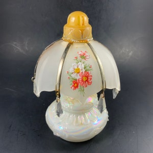 May include: A vintage table lamp with a white, iridescent base and a frosted glass shade. The shade features a floral design with red and white flowers. The lamp has a yellow top and silver metal accents, with hanging crystal-like ornaments.