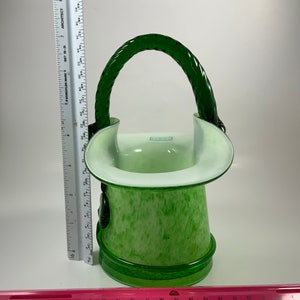Op de afbeelding: Een groen en witte glazen mand met een gevlochten handvat. De mand heeft een gespikkeld patroon op de body en een heldere rand. Het handvat is een donkerdere tint groen. De mand is ongeveer 20 cm hoog.
