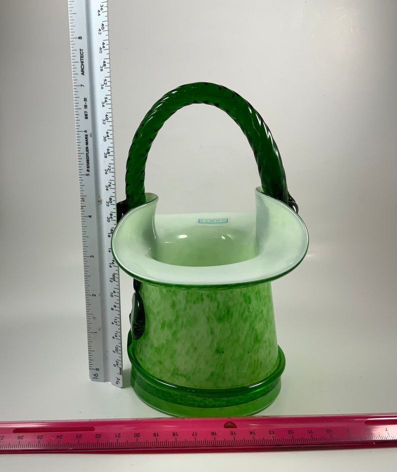 Op de afbeelding: Een groen en witte glazen mand met een gevlochten handvat. De mand heeft een gespikkeld patroon op de body en een heldere rand. Het handvat is een donkerdere tint groen. De mand is ongeveer 20 cm hoog.
