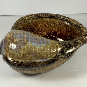 May include: A ceramic seashell-shaped bowl with a textured surface. The bowl features a gradient of brown, tan, and blue hues. The rim has a ridged design, and the interior is a darker brown.