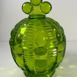 May include: A green glass jar with a lid. The jar has a textured surface with a pattern of circles and lines. The lid is topped with three round, green glass spheres. The jar is a bright, vibrant green color.