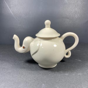 May include: White ceramic teapot shaped like an elephant with a lid and a curled spout. The teapot has a small, red eye painted on the side.
