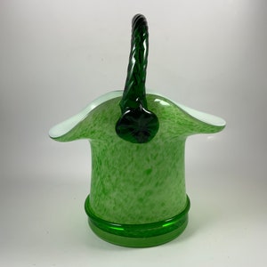 Op de afbeelding: Een groene glazen mand met een witte rand en een gedraaide groene handgreep. De mand heeft een gespikkeld patroon en een effen groene basis. De handgreep is bevestigd aan de mand met een decoratief groen stuk.